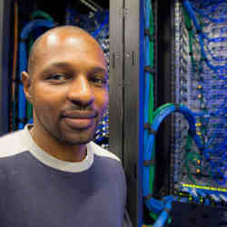 A casually dressed man smiles next to exposed computer components.