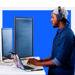 Black man in earphones at laptop computer and two larger displays