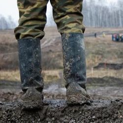 A pair legs with army fatigues stands in a pair of muddy boots in a muddy field.