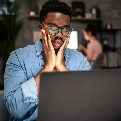 young man holds his face in his hands while looking at a laptop computer