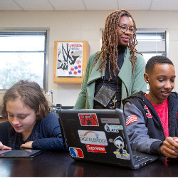 a teacher supervises two students with computers in a classroom