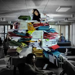 A person sits atop a large mound of paperwork in frustration while others casually go about their day.