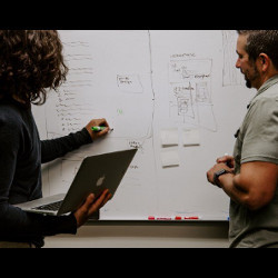 observer watches a person holding a laptop computer and writing on a whiteboard