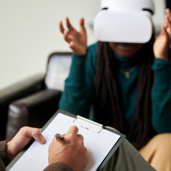 person wearing VR goggles gestures as seated male writes notes