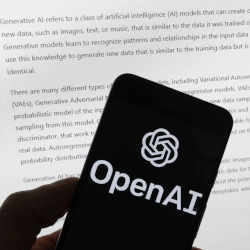 The OpenAI logo on a mobile phone in front of a computer screen displaying output from ChatGPT.